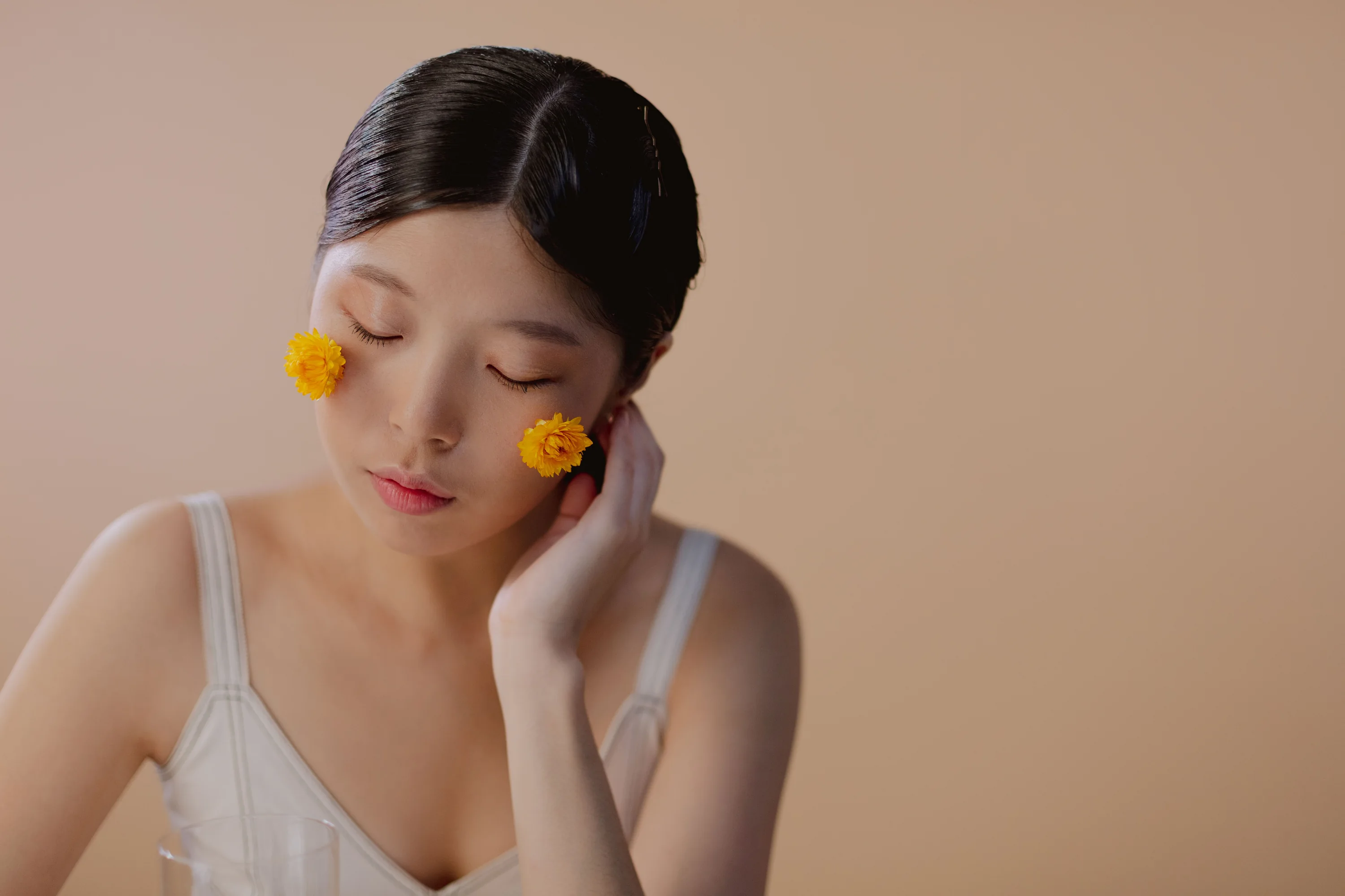 A woman with closed eyes and two small yellow flowers resting on her cheeks against a warm tan backdrop