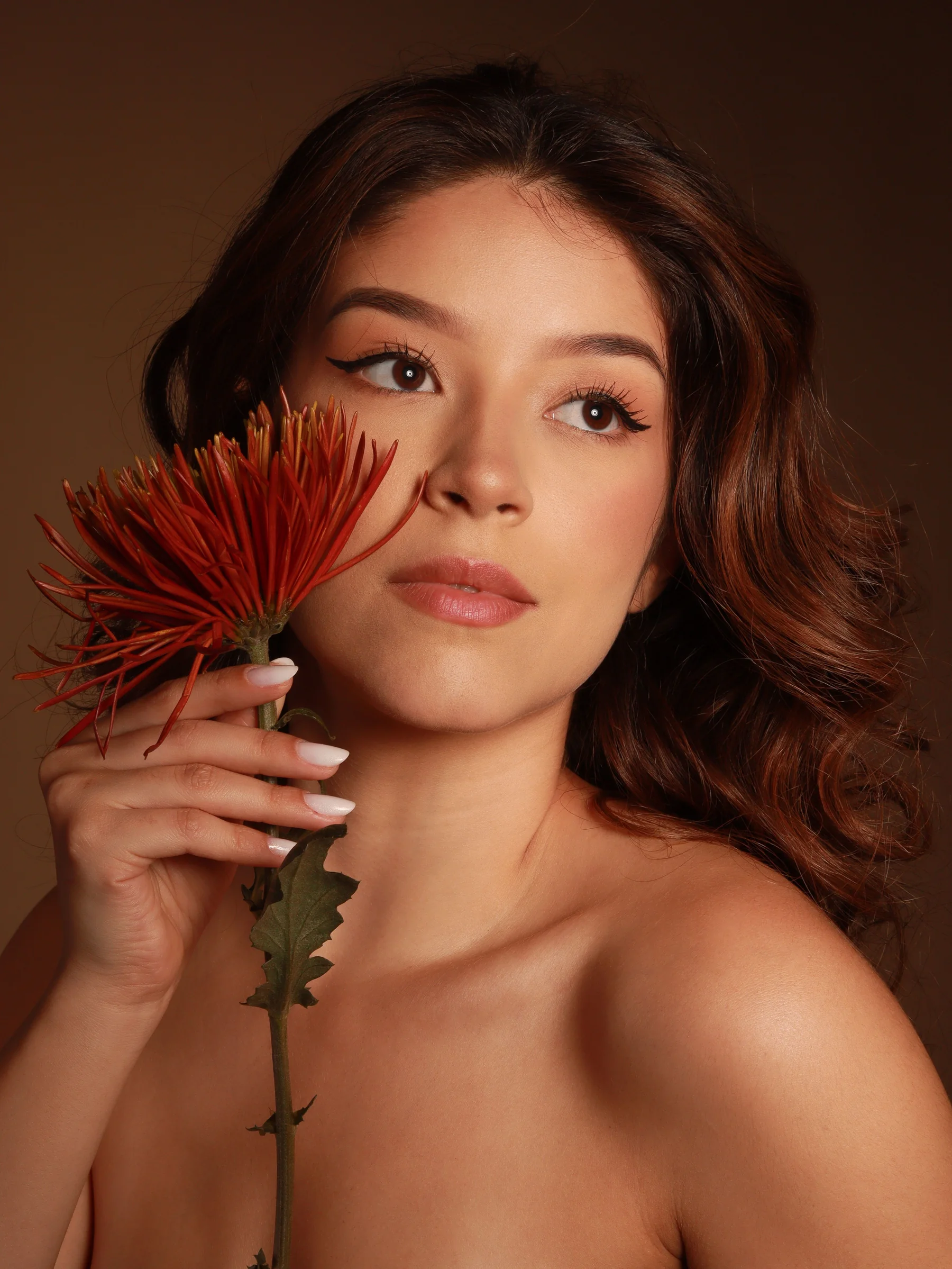 A woman holding a rust-red chrysanthemum beside her cheek against a warm brown backdrop